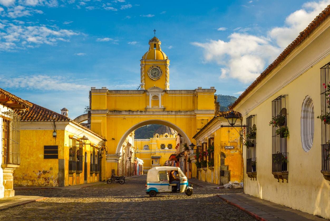 L'arche de Santa Catalina de couleur jaune traversant une rue pavée d'Antigua avec un volcan en arrière-plan